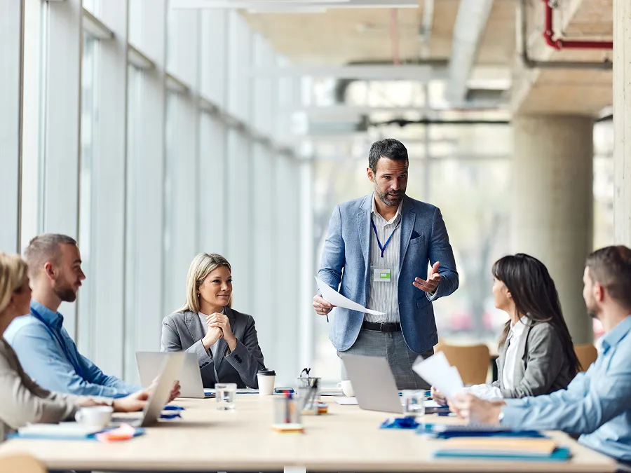 A man stands and speaks to colleagues during a business meeting in a modern office with large windows, discussing L&D Consulting Service Models as other team members listen and look at documents or laptops.