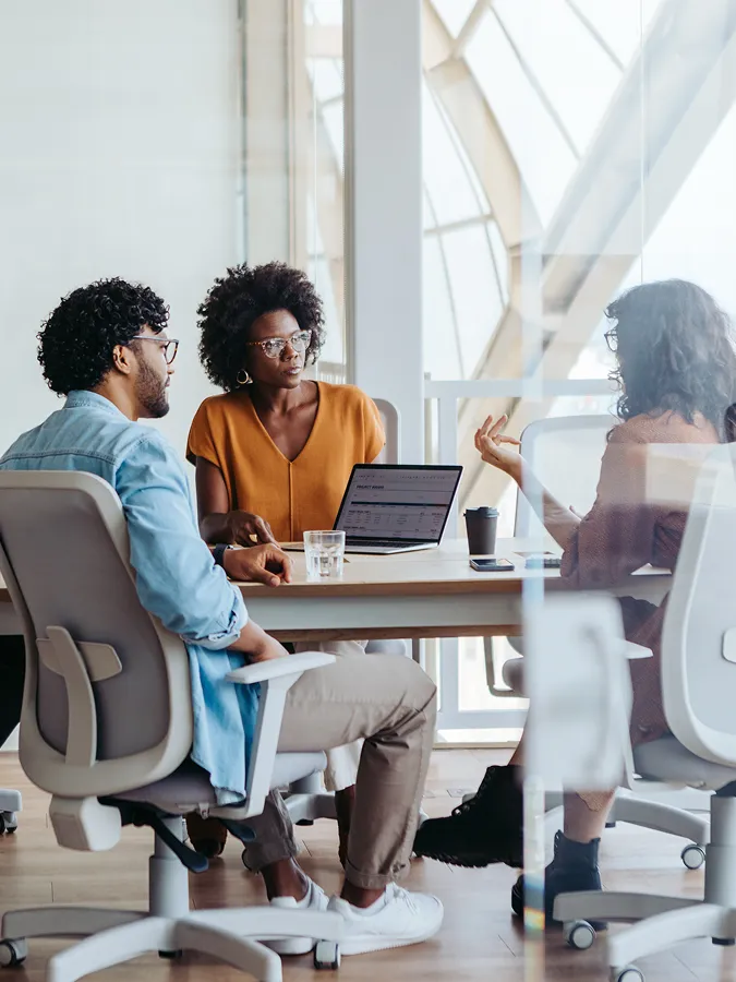 Three people sit around a table in a modern office, having a discussion. A laptop and drinks are on the table. Large windows are in the background.