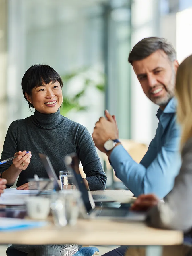 Three people sit at a table in an office, engaged in a discussion with laptops and papers in front of them.