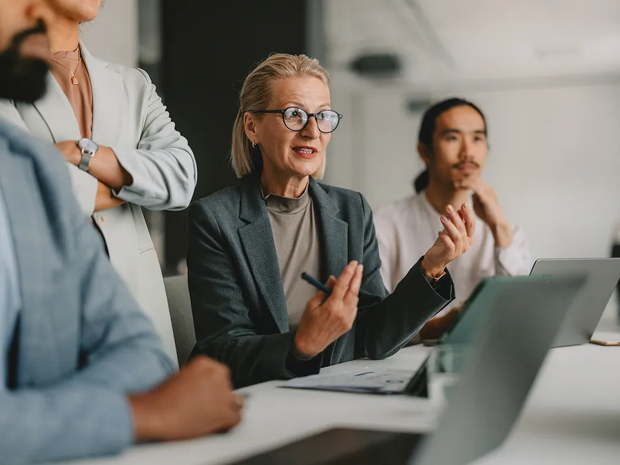 A group of professionals sits at a table in an office meeting, with one woman in glasses speaking while others listen and take notes on laptops.
