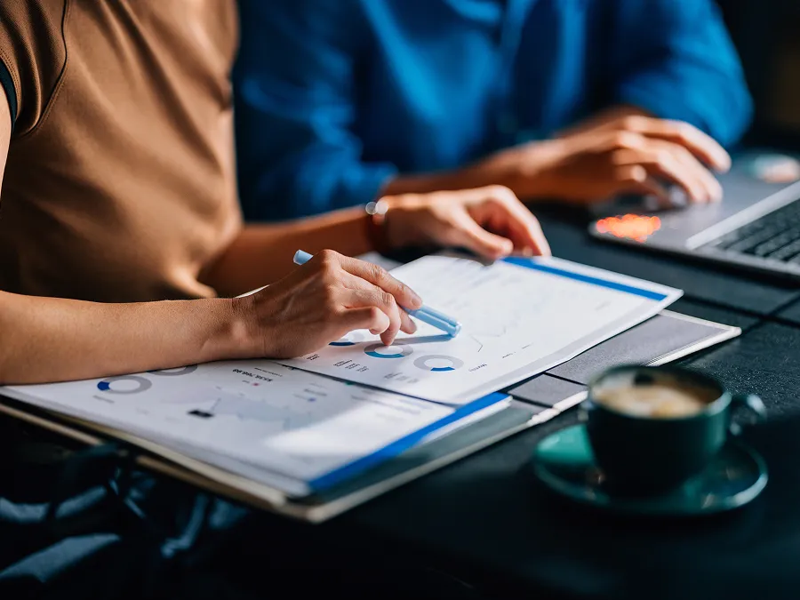 Two people review printed charts and data while working at a table with a laptop and a cup of coffee.