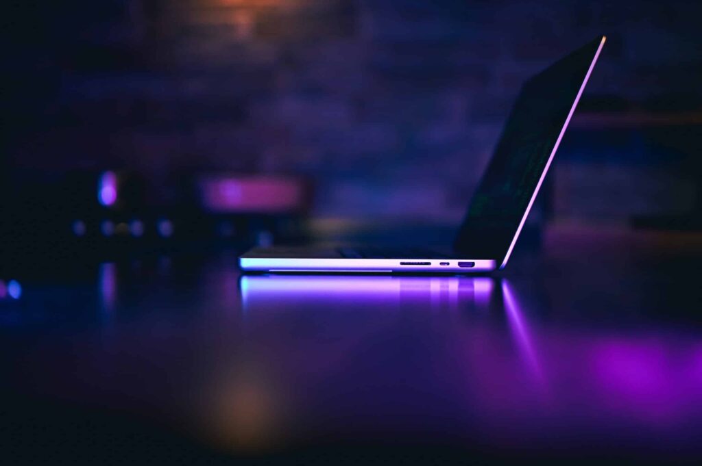 A partially open laptop sits on a reflective surface, illuminated by purple and blue ambient light, with a blurred background.
