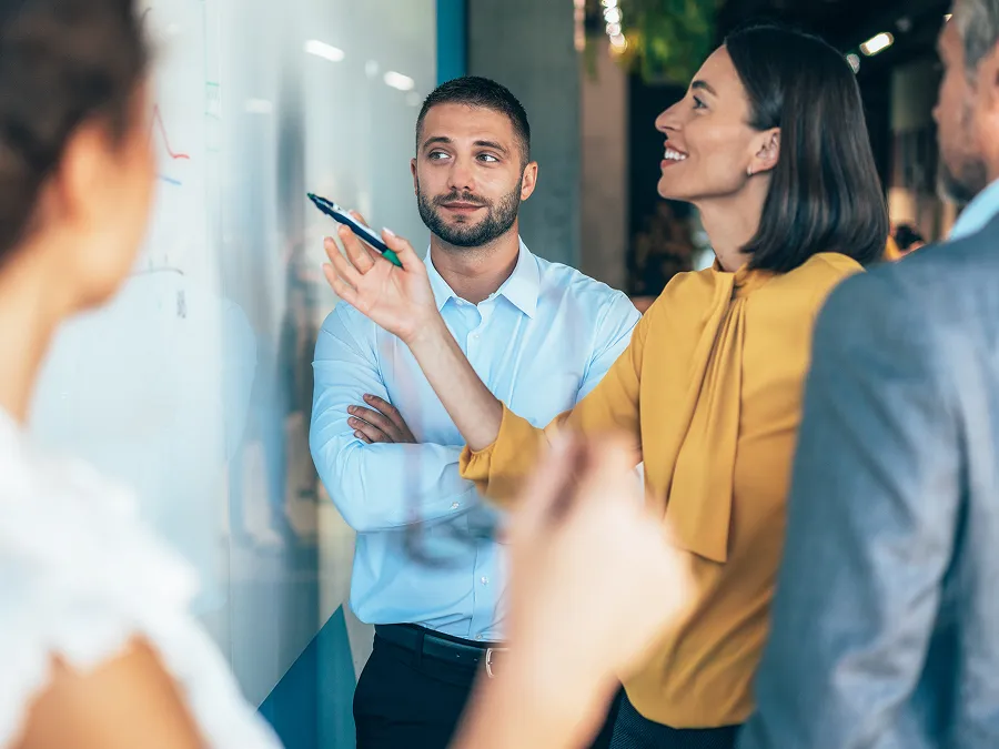 Four people stand around a whiteboard in an office setting. A woman in a yellow blouse points at the board while others watch and listen attentively.