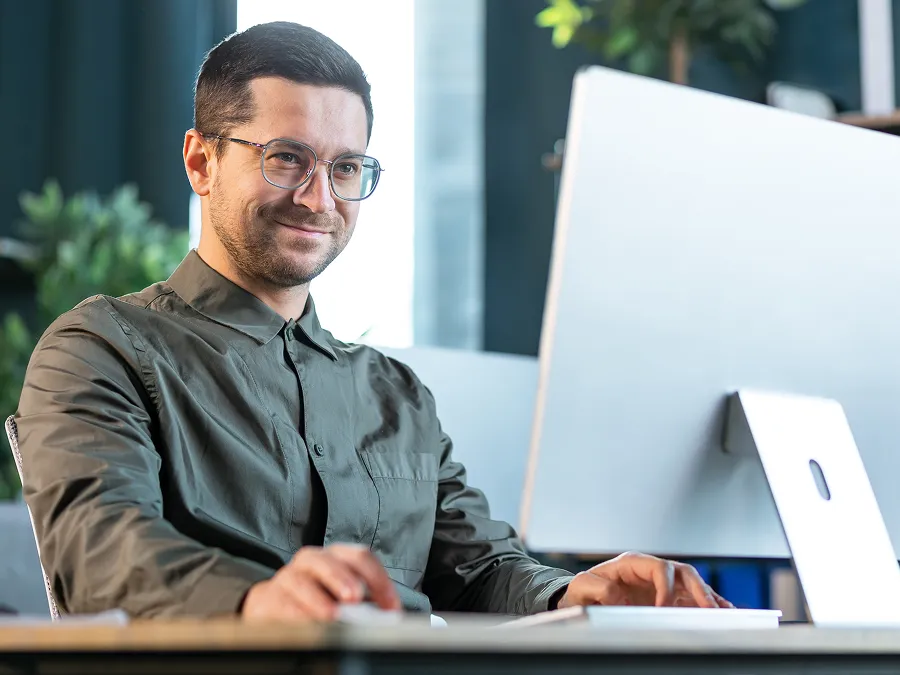 A man wearing glasses and a green shirt is sitting at a desk, using a computer, and smiling slightly.