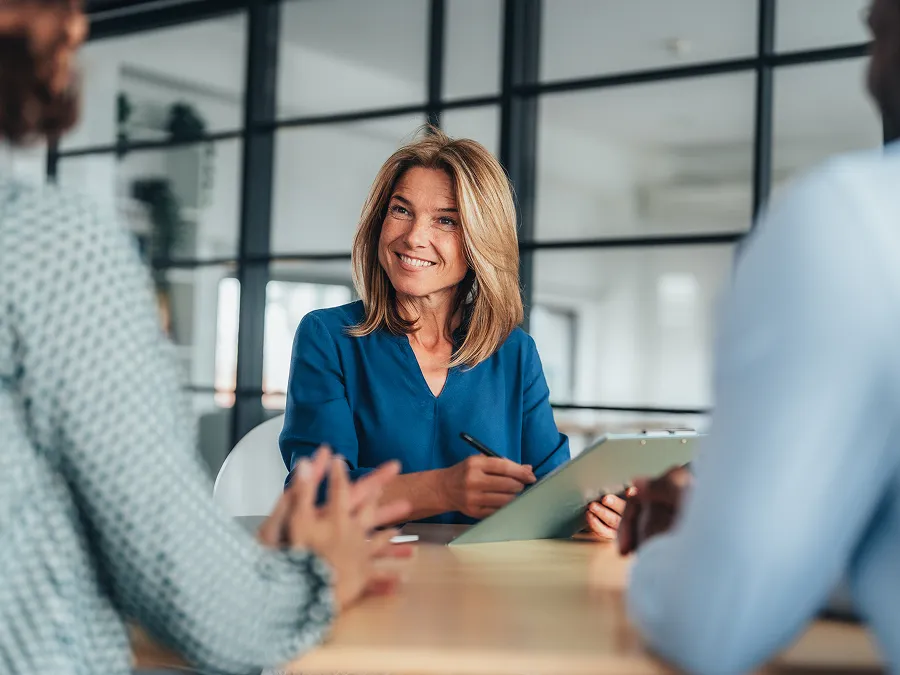 A woman in a blue blouse sits at a desk holding a clipboard, smiling at two people seated across from her in an office setting.
