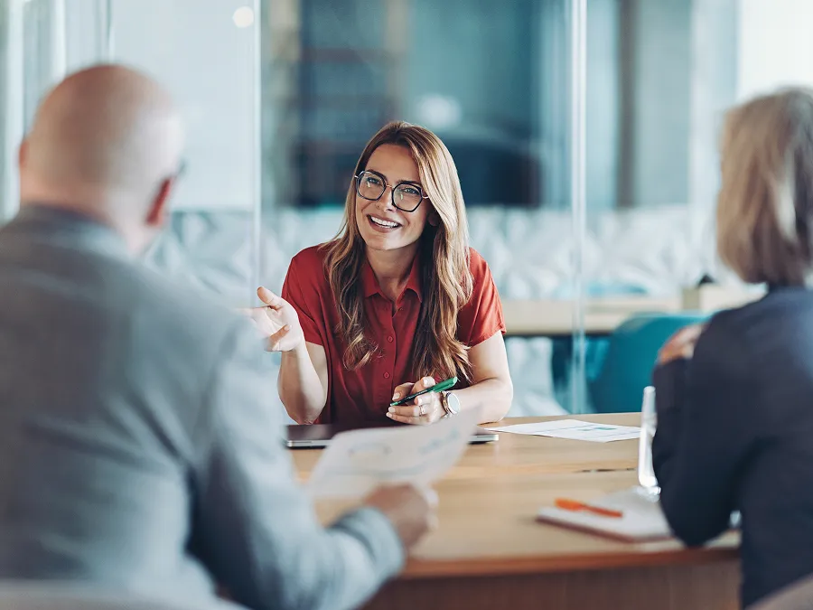 A woman in glasses and a red shirt sits at a table, smiling and talking with two people during a meeting in a modern office setting.