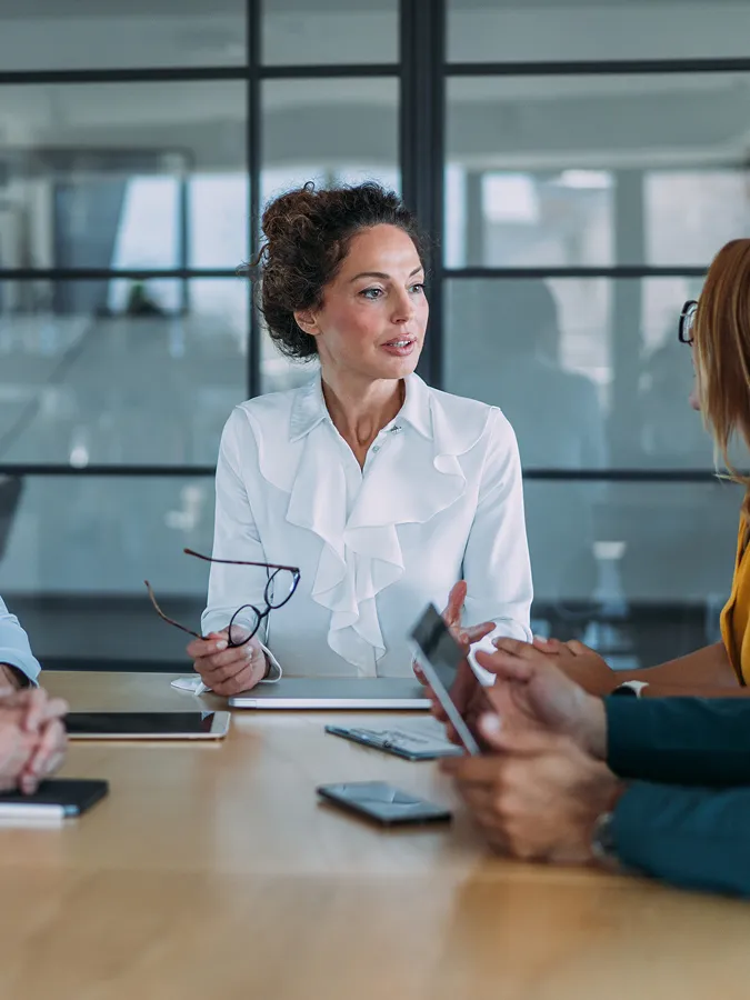 Woman in a white blouse sits at a conference table, holding glasses and speaking to colleagues during a meeting in a modern office.