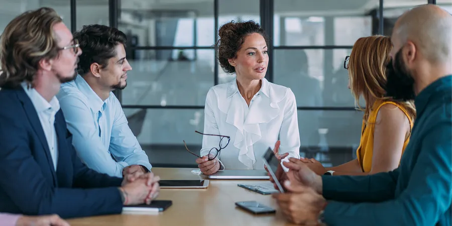A group of six people sit around a conference table in a modern office, having a discussion. One woman in a white blouse appears to be leading the meeting.