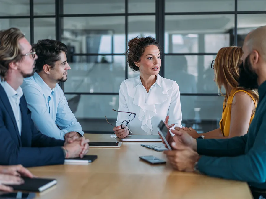 A woman leads a business meeting with five colleagues seated around a conference table in a modern office setting.