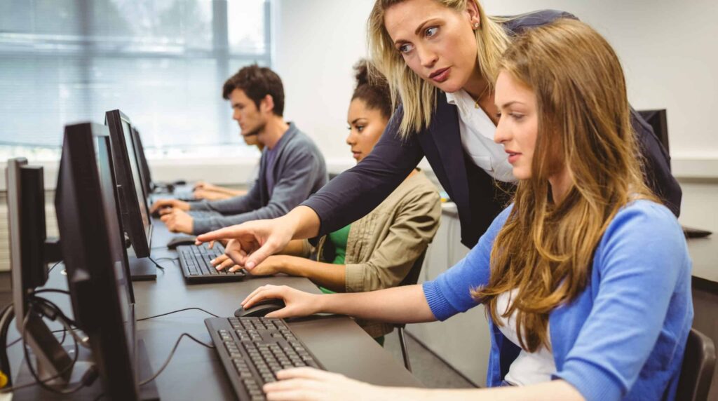 A teacher assists a student at a computer while other students work at their own computers in a classroom.