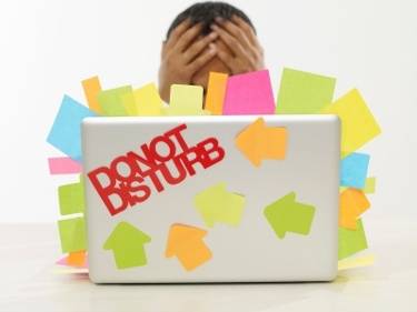 A person sits behind a laptop covered in colorful sticky notes and a red "DO NOT DISTURB" sign, with their head in their hands.