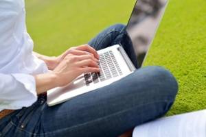 Person sitting cross-legged on grass, typing on a laptop. Only hands, lap, and part of the torso are visible. An open book rests nearby.