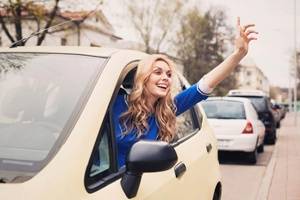 Woman leaning out of a car window, smiling and waving, on a residential street with parked cars and trees in the background.