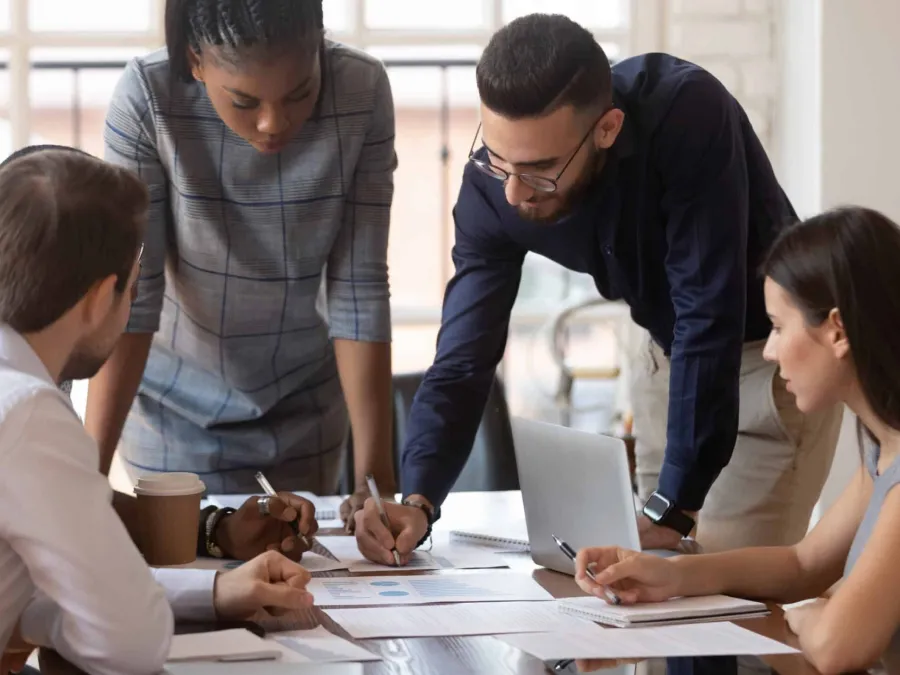 Four people sit and stand around a table, reviewing documents and charts together in an office meeting setting. A laptop and coffee cup are visible on the table.