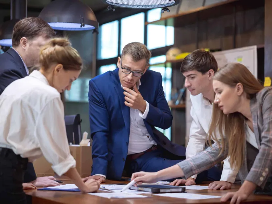 Five people in business attire stand around a table in an office, looking at documents and discussing work.