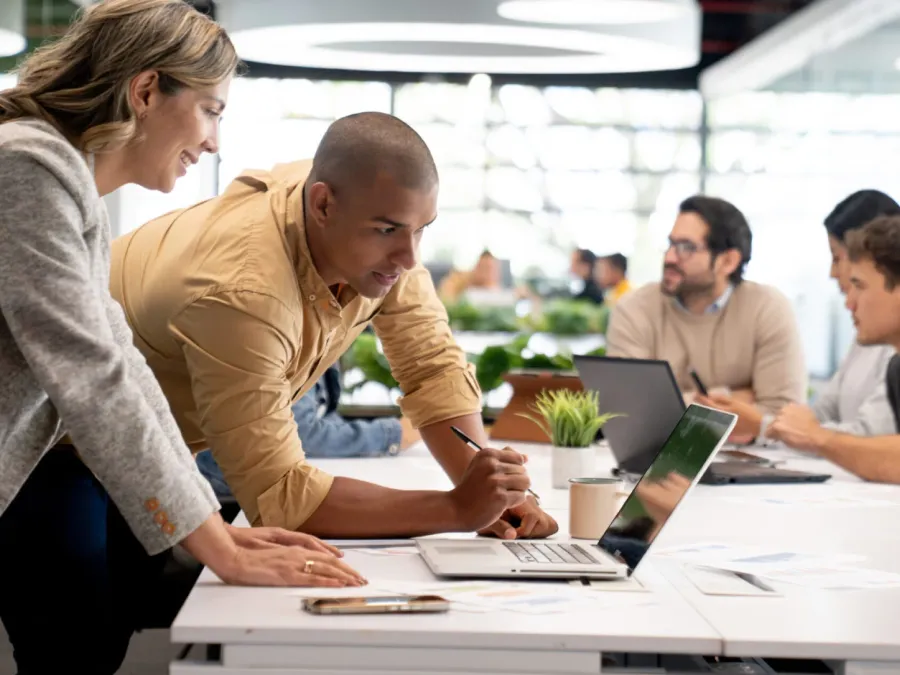 Two people collaborate at a laptop while two others have a discussion in the background in a modern office setting.