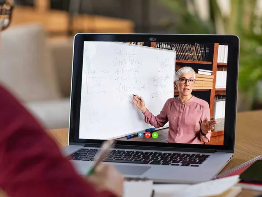 Person watching an online lecture; woman stands by a whiteboard covered in equations, explaining concepts via video call on a laptop.