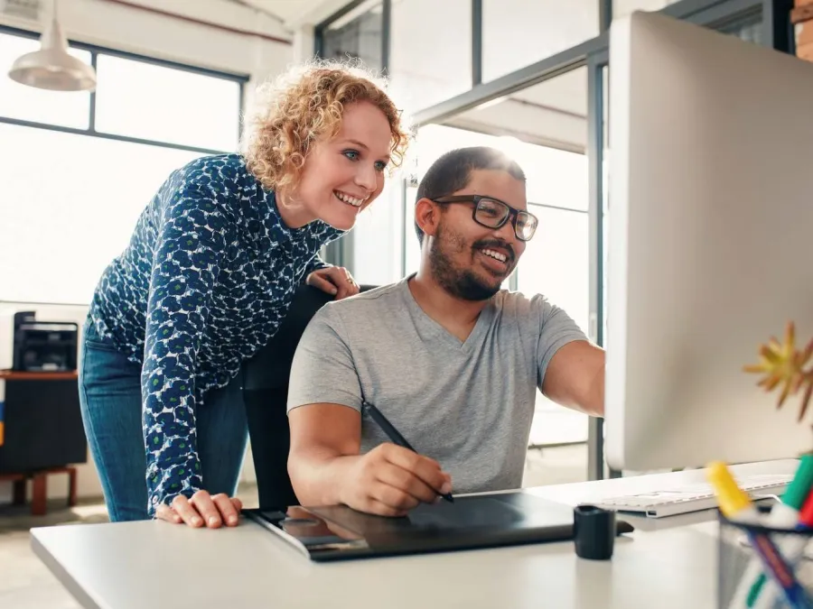 A man sitting at a desk works on a computer while a woman stands beside him, both looking at the screen and smiling in a modern office setting.
