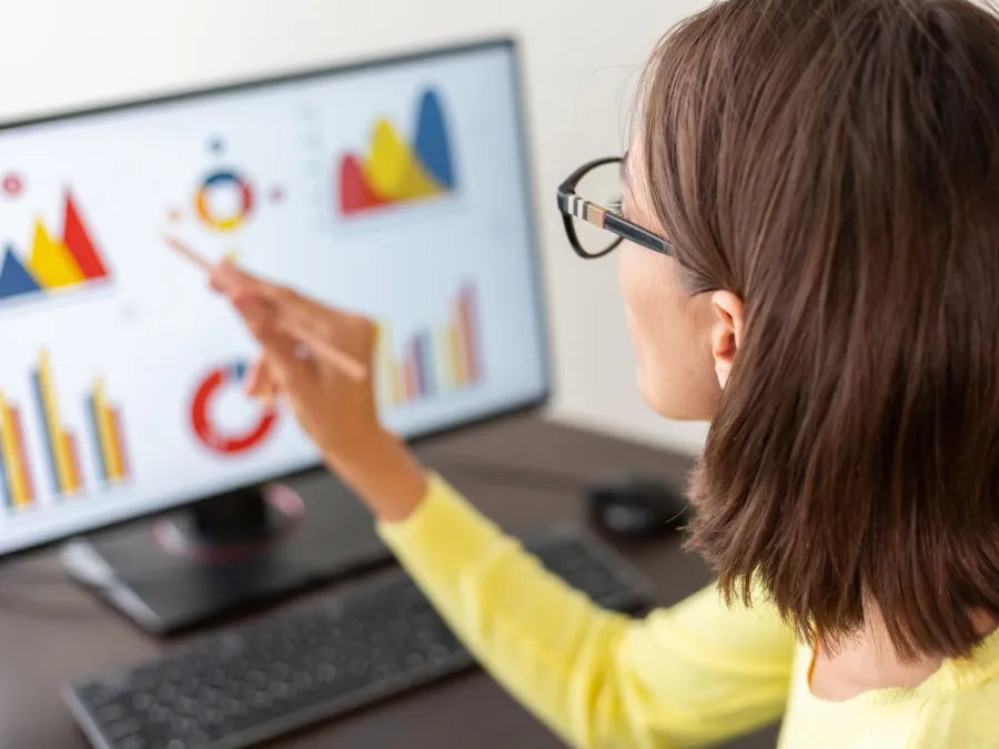 A woman wearing glasses points at colorful charts and graphs displayed on a computer monitor at her desk.