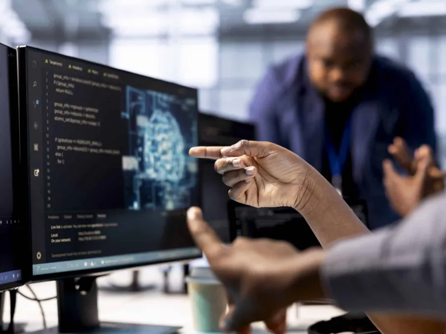 Two people discuss code displayed on a computer monitor, with one person pointing at the screen and blurred office environment in the background.
