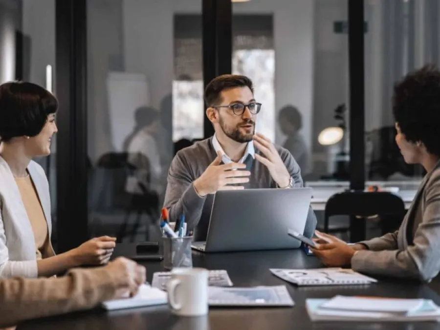 Four people sit around a table in an office, discussing work. One man gestures while speaking, with a laptop in front of him. Documents, mugs, and pens are on the table.