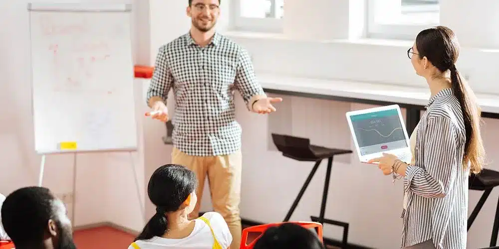 A man speaks to a seated group in a bright room while a woman holding a laptop stands nearby; a chart is visible on a whiteboard in the background.