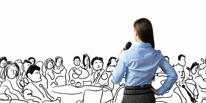 A woman holding a microphone stands facing a large audience, which is illustrated as a line drawing on a white background.