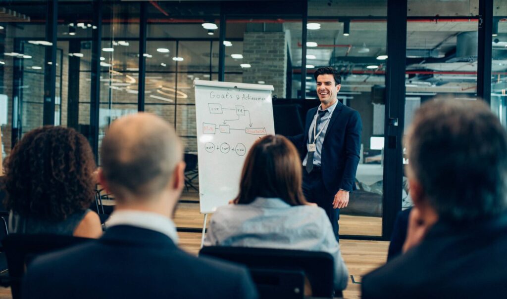 A man in a suit presents business strategies on a flip chart to a seated audience in a modern office meeting room.