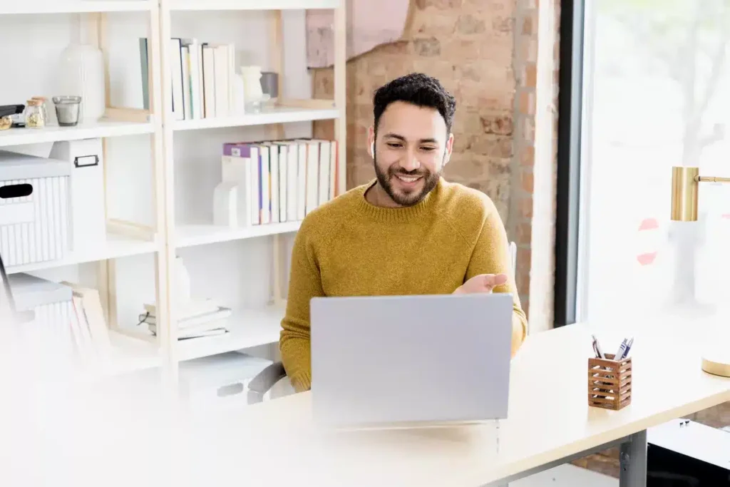 A man sits at a desk in a modern office, smiling and gesturing while looking at a laptop screen. Bookshelves and a window are visible in the background.