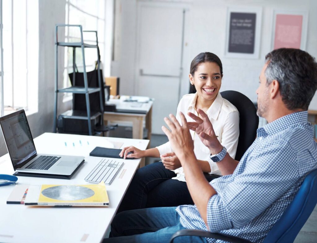 Two people sit at a desk in an office, talking and smiling. A laptop, notebook, and papers are on the desk. Shelves and framed prints are in the background.