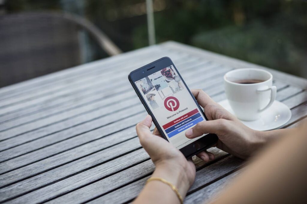 Person holding a smartphone with the Pinterest login screen open, sitting at a wooden table with a white cup of coffee.