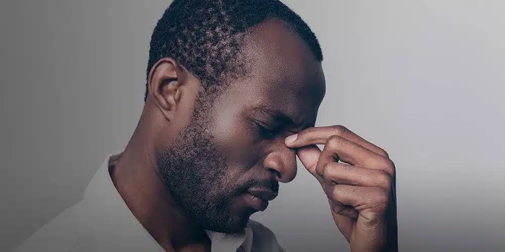 A man with a beard presses his fingers to the bridge of his nose, eyes closed, appearing tired or stressed, against a plain background.