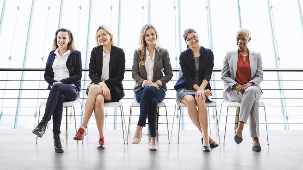 Five women in business attire sit on chairs in a row, facing the camera, in a modern, well-lit office setting with large windows in the background.