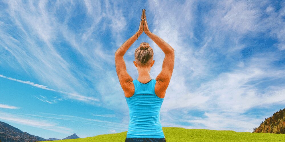 A person with blond hair in a blue tank top stands outdoors with arms raised overhead in a yoga pose, set against a grassy field and a partly cloudy sky.