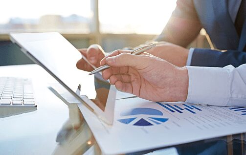Two people in business attire review charts and graphs on paper and a tablet at a desk in an office setting.