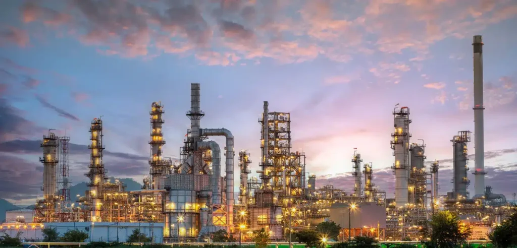 Industrial oil refinery with multiple towers and pipes, illuminated at dusk against a cloudy sky.