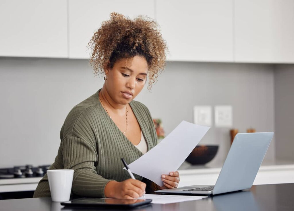 A woman sits at a kitchen counter with a laptop, holding papers in one hand and writing with the other. A coffee mug and tablet are also on the counter.