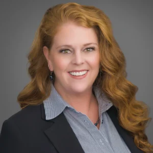 A woman with long, wavy red hair wearing a dark blazer and striped shirt smiles at the camera against a gray background.