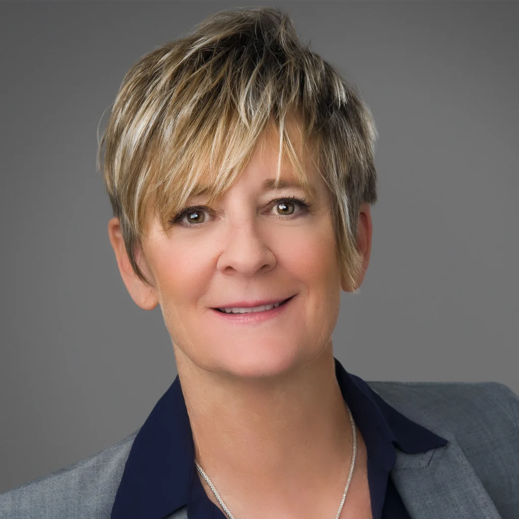 A person with short, blonde hair wearing a gray blazer over a dark shirt, smiling slightly, poses in front of a plain gray background.