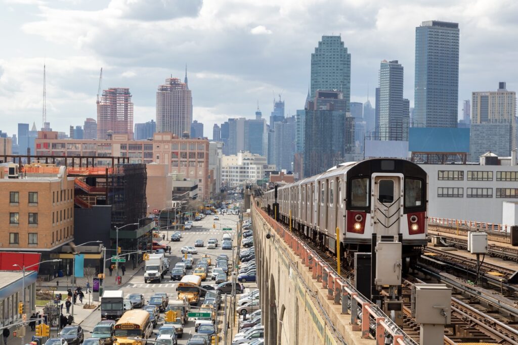 A subway train travels on an elevated track above a busy street with traffic, set against a city skyline with tall buildings under a cloudy sky.