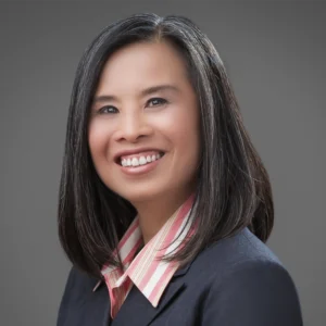 A woman with straight black hair, wearing a navy blazer and a pink striped collared shirt, smiles in a professional studio portrait against a gray background.