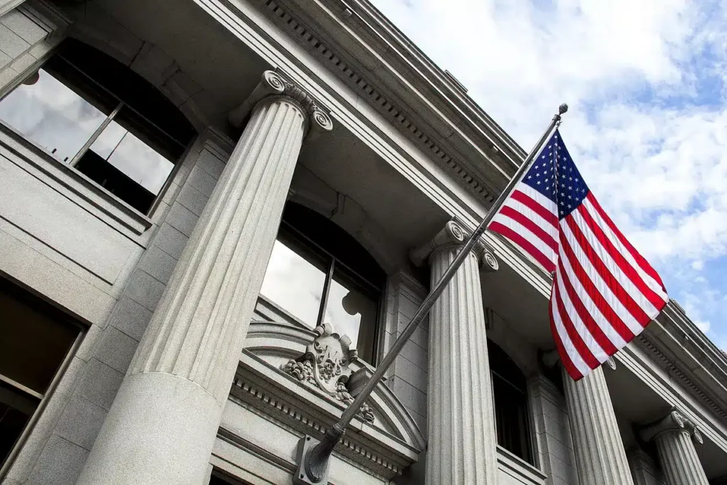 A U.S. flag on a pole is displayed outside a neoclassical building with large columns and decorative stonework.