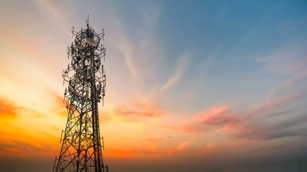 A telecommunications tower with multiple antennas stands against a colorful sunset sky with scattered clouds.
