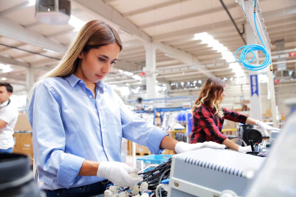 Two women wearing gloves work on assembling electronic equipment at a workstation in a brightly lit industrial facility.