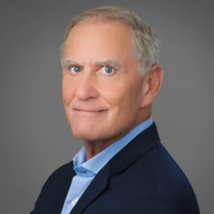 A middle-aged man with short gray hair, blue eyes, and a slight smile, wearing a blue blazer and light blue shirt, posed against a plain gray background.