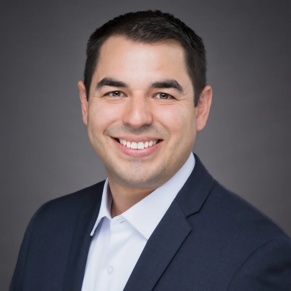 A man in a dark suit jacket and white shirt smiles at the camera against a plain gray background.