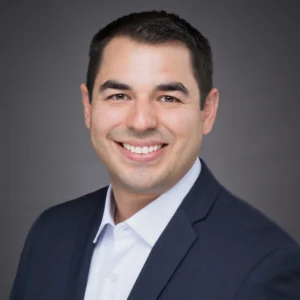 A man in a dark suit jacket and white shirt smiles at the camera against a plain gray background.