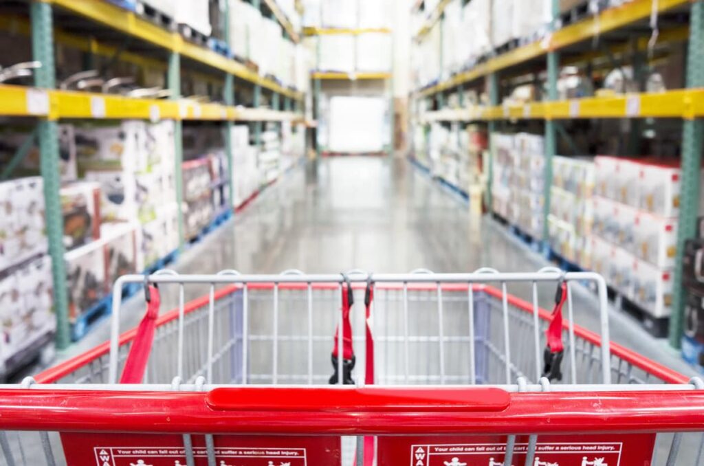 A shopping cart is pushed down an aisle in a warehouse-style store, surrounded by stocked shelves and lit by bright overhead lights.