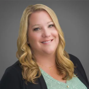 A woman with long blonde hair wearing a green polka dot blouse and dark blazer, smiling in front of a gray background.