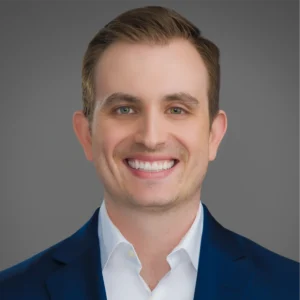 A man in a blue suit jacket and white shirt smiles at the camera against a plain gray background.
